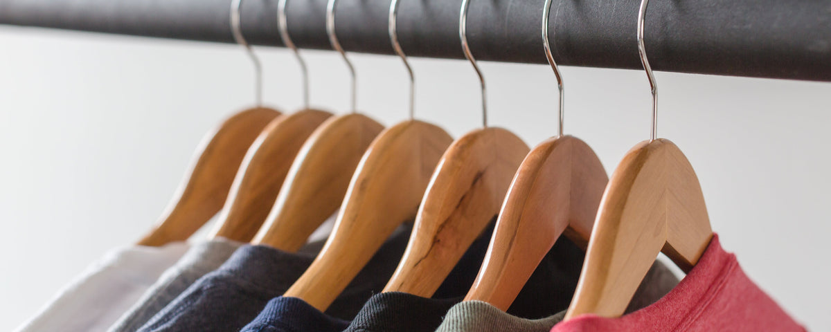 A close-up of several wooden hangers hanging from a black clothing rack, with the tops of different garments partially visible in soft focus.